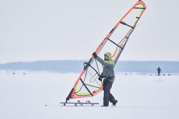 A middle-aged man, a snowsurfer, rolls out a sailboard onto a snow-covered frozen lake. Preparing for snowsurfing on a cloudy winter day.