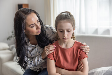 Mother comforts upset daughter that sits with arms crossed on a couch. - 590511731