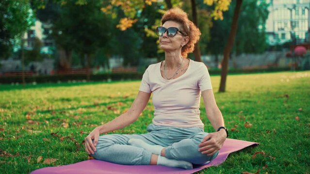 Senior woman meditating and exercising yoga lotus position outdoors. Ederly female doing stretching exercises on yoga mat in park on green lawn at sunset. Feeling energy. Concept calm and meditation.