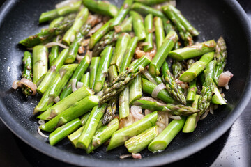 Green asparagus pieces with onion in a black frying pan, cooking concept for vegetarian vegetable meal with spring time vegetables, selected focus