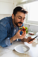 Close-up shot of a handsome young man eating a sandwich for breakfast, while holding a phone.
