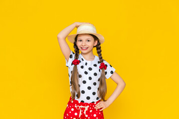 A happy little girl is holding onto a straw hat and smiling broadly, enjoying the bright summer. A teenage girl in polka dot clothes and two pigtails on a yellow isolated background.