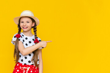 A beautiful little girl in polka dot shorts and a straw hat is pointing at your advertisement. Summer holidays. A teenage girl is waiting for summer on a yellow isolated background. Copy Space