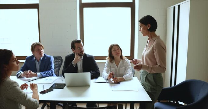 Mature female business leader talking to employees at meeting table, standing, speaking, instructing workers. Mentor woman explaining work project tasks to interns