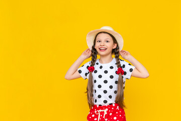 A happy little girl is holding onto a straw hat and smiling broadly, enjoying the bright summer. A teenage girl in polka dot clothes and two pigtails on a yellow isolated background.