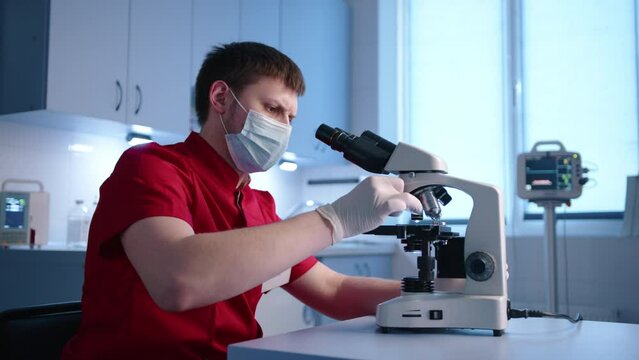 Young male serious animal doctor checks samples for analysis using binocular microscope. The vet has red doctor suit, latex medical gloves and medical mask. High quality 4k footage