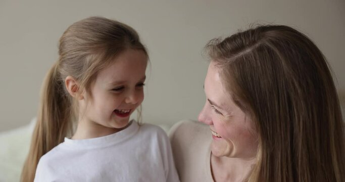 Happy mom and joyful little daughter kid hugging, smiling, laughing, looking at camera, sitting close on bed. Cheerful mother talking to cute girl, enjoying motherhood, family leisure