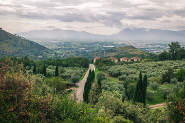 A road winding through a lush green valley with mountains in the background and a cloudy sky above it, Lazio, Italy