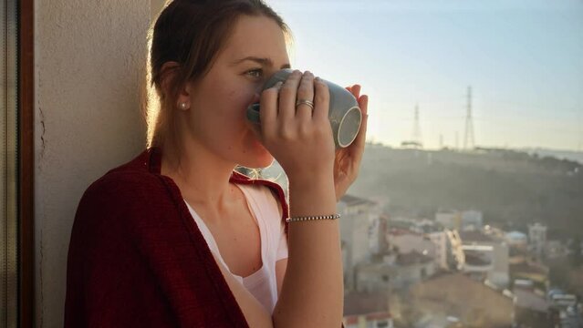 Portrait of beautiful woman standing on balcony with coffee and looking on sunrise at city. People relaxing, resting at home, beautiful cityscape