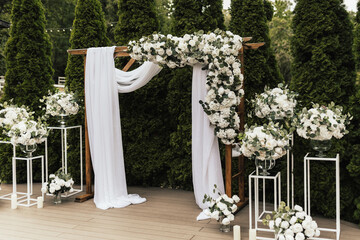 A beautiful wooden wedding arch decorated with white flowers and greenery, stands in the garden against the background of tui trees.