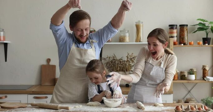 Happy family couple of parents and little daughter baking in kitchen together, having fun, throwing raining sifting flour on table dish, eggs, pastry ingredients, rolling dough, cooking dessert