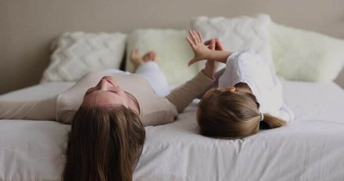 Relaxed playful mom lying on back on bed, tickling, cuddling, catching kid, playing with child, pretending sleepy, enjoying leisure, relaxation, active games, motherhood, having fun