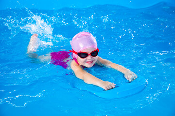 Smiling little girl learning to swim in indoor pool with flutterboard during swimming class