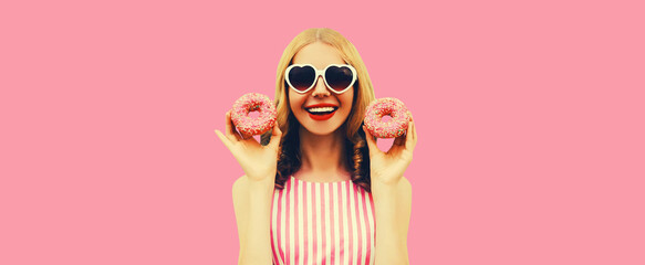 Portrait of happy cheerful laughing young woman with two sweet donuts having fun wearing heart shaped sunglasses on pink background