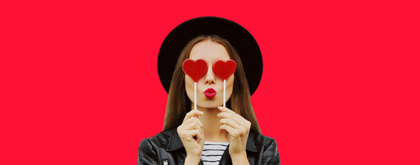 Portrait of beautiful young woman covering her eyes with red heart shaped lollipop blowing lipstick...