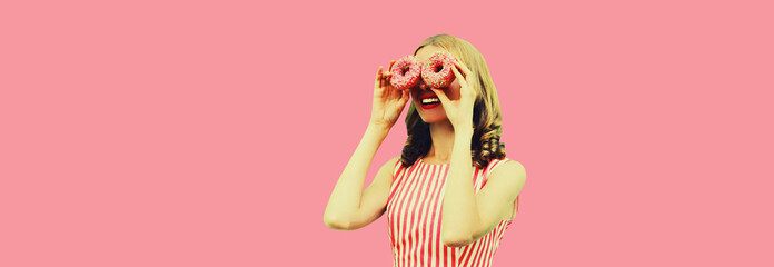 Portrait of happy funny smiling woman covering her eyes looking for something looking through donuts as binoculars having fun on pink background