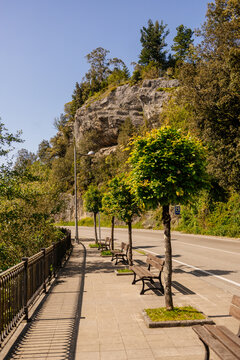 A city path with benches in a park with a rock. The rock above the road is protected by a net to protect people from falling stones