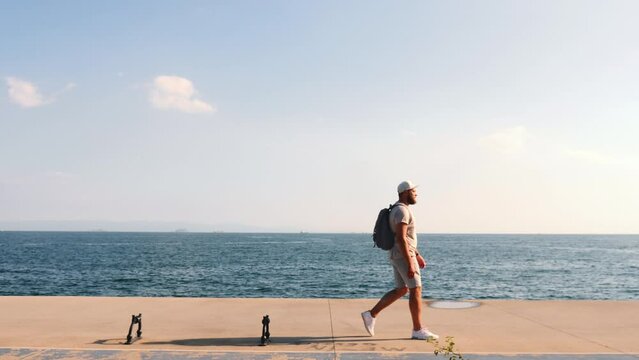 Happy young tourist man walking parallel to camera alone alongside sea coast on a sunny day, enjoying sea side