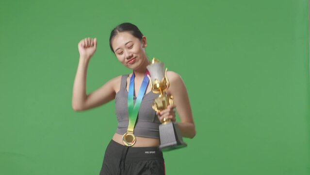 Asian Woman With A Gold Medal Looking At A Gold Trophy Then Celebrating Winning As The First Winner On Green Screen Background In The Studio
