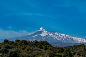 Wide-angle shot of snow-covered Etna as it erupts white puffs of steam against an intensely blue sky