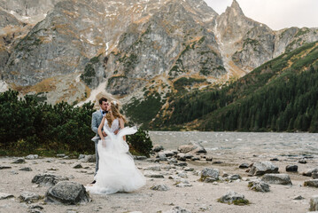 Romantic wedding couple in love standing on the stony shore of the Sea Eye lake in Poland. Scenic mountain view. The bride and groom. Morskie Oko. Tatra mountains.