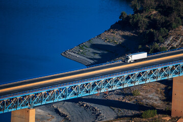 Truck with a refrigerated trailer driving on a metal bridge over a blue water swamp.