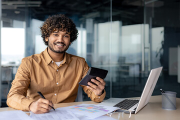 Portrait of young successful financier accountant at work inside office building at workplace, hispanic man smiling and looking at camera, man using calculator in paperwork.