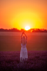 Faceless young blonde woman stands in purple field at sunset in bright rays of the sun. Travel in...