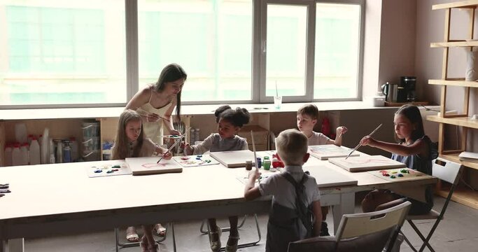 Diverse group of little kids painting in acrylic colors on drawing class, studying creativity in artistic school. Young female teacher training kids in creativity, checking canvas
