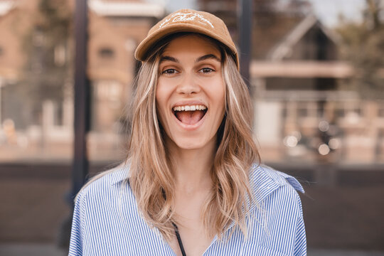 Cute young woman with a lovely sense of humor standing leaning against a glass window exterior with copy space in an urban street laughing at the camera. Attractive blonde girl wear shirt and cap.
