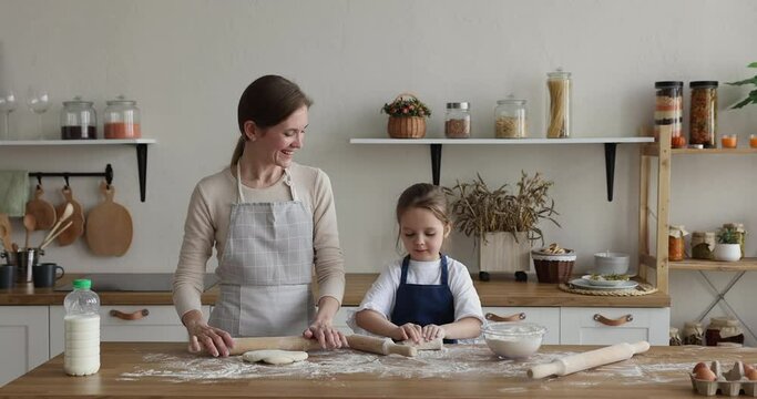 Engaged young mom and sweet little kid girl in aprons rolling dough on kitchen table with flour, baking pie, using rollers. Mother teaching child to cook traditional pastry, preparing dessert