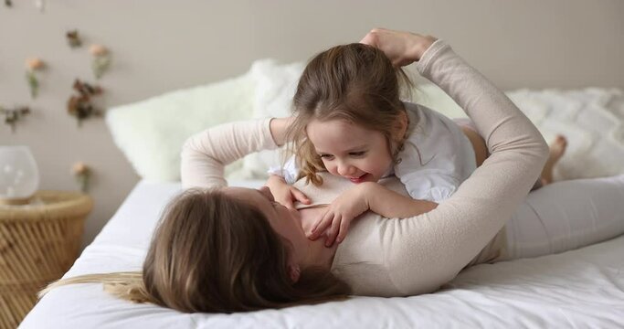 Happy cute little kid girl resting on playful mom, laughing, having fun. Cheerful mother lying on back on bed, cuddling sweet child, holding Daughters hands, shaking head, playing active games