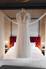 A perfect wedding dress with a lush skirt on a hanger, next to a bed with a red headboard and a white sheet, and a sconce that illuminates the bride's room.
