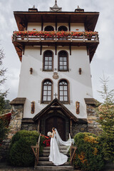 Romantic full-length portrait of a brown-haired woman in a white dress with a bouquet standing near the wooden gate of the church entrance