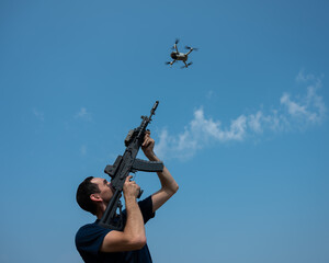 A man aims to shoot a rifle at a flying drone against a blue sky. 