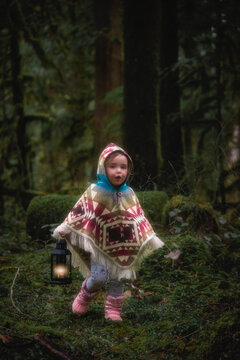 Little toddler girl exploring in the dark lush Pacific Northwest forest carrying lantern wearing poncho
