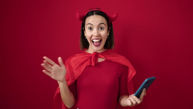 Young beautiful hispanic woman wearing devil costume using smartphone over isolated red background