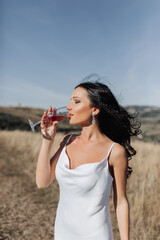 A beautiful girl with closed eyes and beautiful black hair in a white dress is drinking pink champagne. Beautiful contours of the face. Portrait of a girl. Earrings