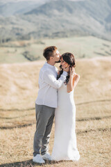 A stylish model couple in the mountains in the summer. A young boy and a girl in a white silk dress are walking on the slope against the background of the forest and mountain peaks. boy kisses a girl