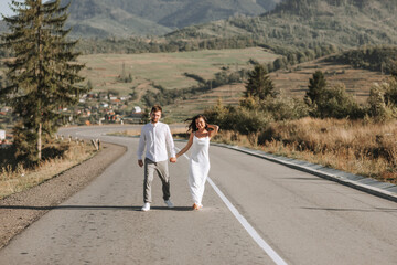 A stylish groom in a white shirt and a cute brunette bride in a white dress are walking on an asphalt road against the background of a forest and mountains. Wedding portrait of newlyweds.