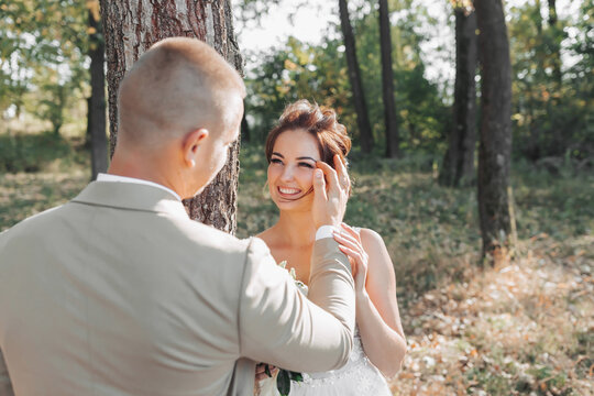 Wedding photo in nature. The bride and groom are standing near a tree. The bride looks at the groom, and he gently fixes her curls. Meeting in the forest.