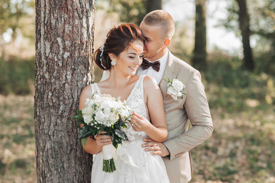 Wedding photo in nature. The bride and groom are standing near a tree, the groom hugs his beloved from behind and kisses her, she smiles sincerely. Portrait. Summer wedding