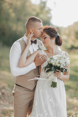 Wedding photo in nature. The bride and groom are standing in the forest, the groom hugs his beloved from behind and kisses her, she smiles sincerely. Portrait. Summer wedding