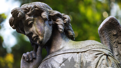 Obraz premium Contemplative Angel Statue with Leaves features a camera view up at an angel statue with leaf shadows on the body and defocused green leaves in the background.