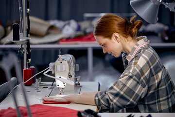 seamstress lady in casual wear sit behind table using sewing machine, making modern clothes, young redhead caucasian woman at workshop, holding red fabric, enjoy tailoring, concentrated