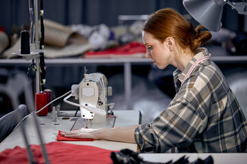 seamstress woman in casual wear sit behind table using sewing machine, making modern clothes, young redhead caucasian female at workshop, holding red fabric, enjoy tailoring, concentrated