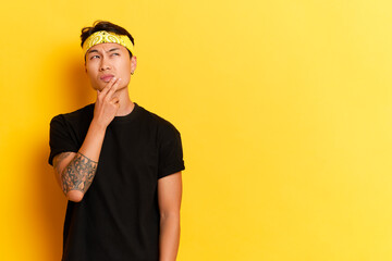 Portrait of thoughtful asian man in black t-shirt and yellow bandana posing on yellow studio background, does a spot of thinking at some point, copy space, high quality photo