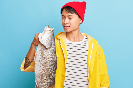 Asian boy holds big fish in his hand, wearing red cap yellow jacket, looks at his catch with satisfaction, good luck concept, copy space, high quality photo