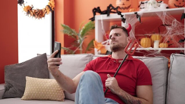 Young caucasian man wearing devil costume sitting on the sofa using smartphone at home