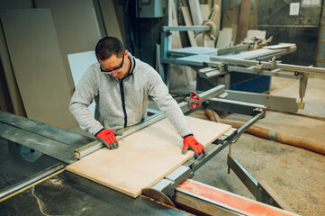 A woodworker is cutting wooden panel on a saw in his workshop.